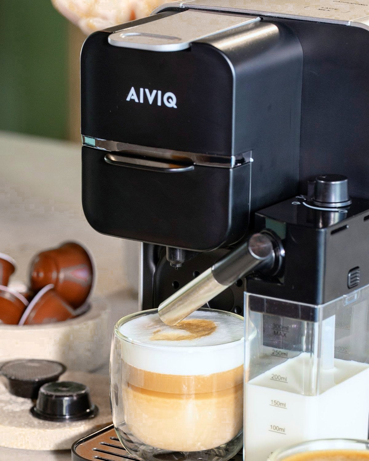 AIVIQ MultiBrew Pro mit einem Latte macchiato, Kapseln und dem Milchbehälter auf einem Steintisch